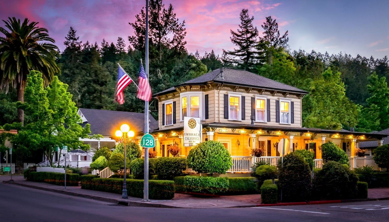 The quaint Embrace Calistoga is pictured under a pink sunset sky, surrounded by trees on a street corner | Best Calistoga Hotels