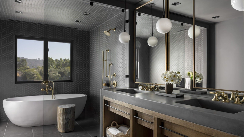 A modern luxury bathroom with dark tiled walls, a dark vanity with gold hardware, and a large white tub underneath a window | Best Calistoga Hotels