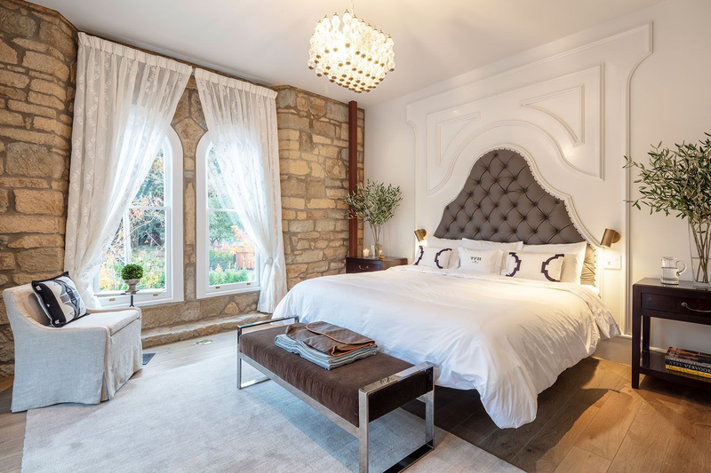 A spacious bedroom with a large white bed against a white wall and a stone wall off to the side with a large window and white curtains | Best Calistoga Hotels