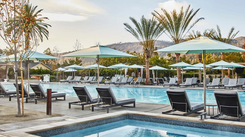 A long, rectangular pool is surrounded by pool chairs and umbrellas, with palm trees scattered around | Best Calistoga Hotels