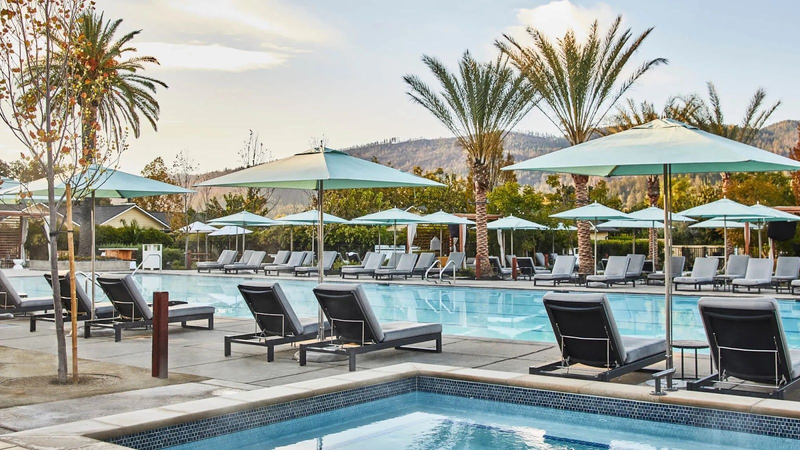 A long, rectangular pool is surrounded by pool chairs and umbrellas, with palm trees scattered around | Best Calistoga Hotels